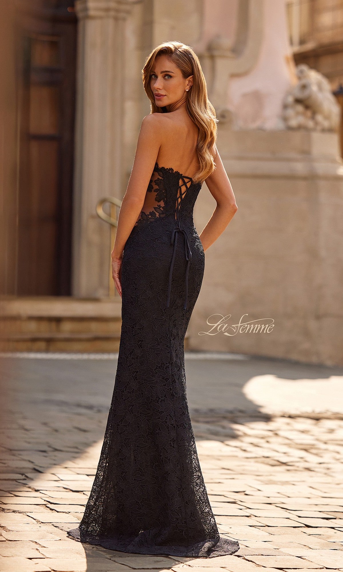 Wearing the La Femme 33316 long fitted strapless lace prom dress with a lace-up back and sweetheart neckline, a woman stands on a cobblestone street, looking over her shoulder.
